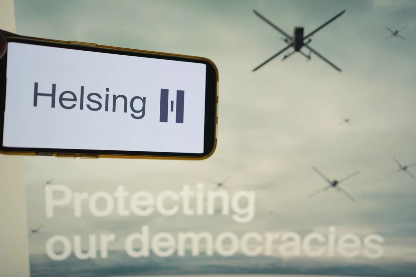 Phone screen displaying the Helsing logo against a backdrop of military drones and the company's "Protecting our democracies" tagline. The Munich-based defence AI company is the largest private AI company in Europe, valued at $14.1 billion as of June 2025.